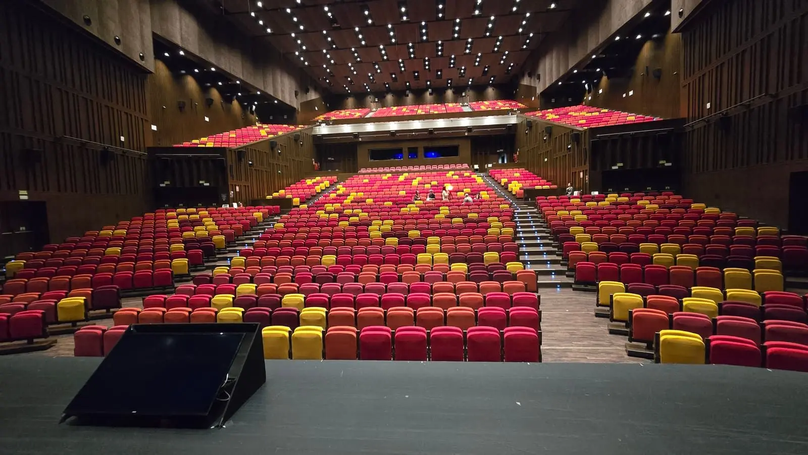 Auditorio vacío visto desde el escenario, con cientos de butacas rojas y amarillas en gradas