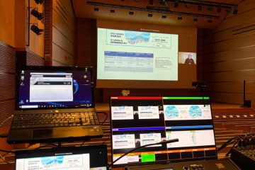 Puesto de control audiovisual con dos portátiles y una mesa de mezclas, con la sala y la pantalla al fondo.
