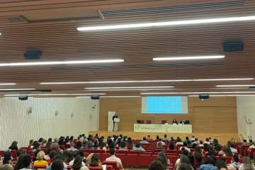 Sala de conferencias llena de público sentado en butacas rojas, con el ponente al fondo bajo una pantalla.
