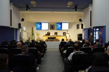 Sala de actos llena de público visto desde el fondo, con la mesa de ponentes y dos pantallas al frente.
