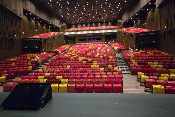 Auditorio vacío visto desde el escenario, con cientos de butacas rojas y amarillas en gradas.