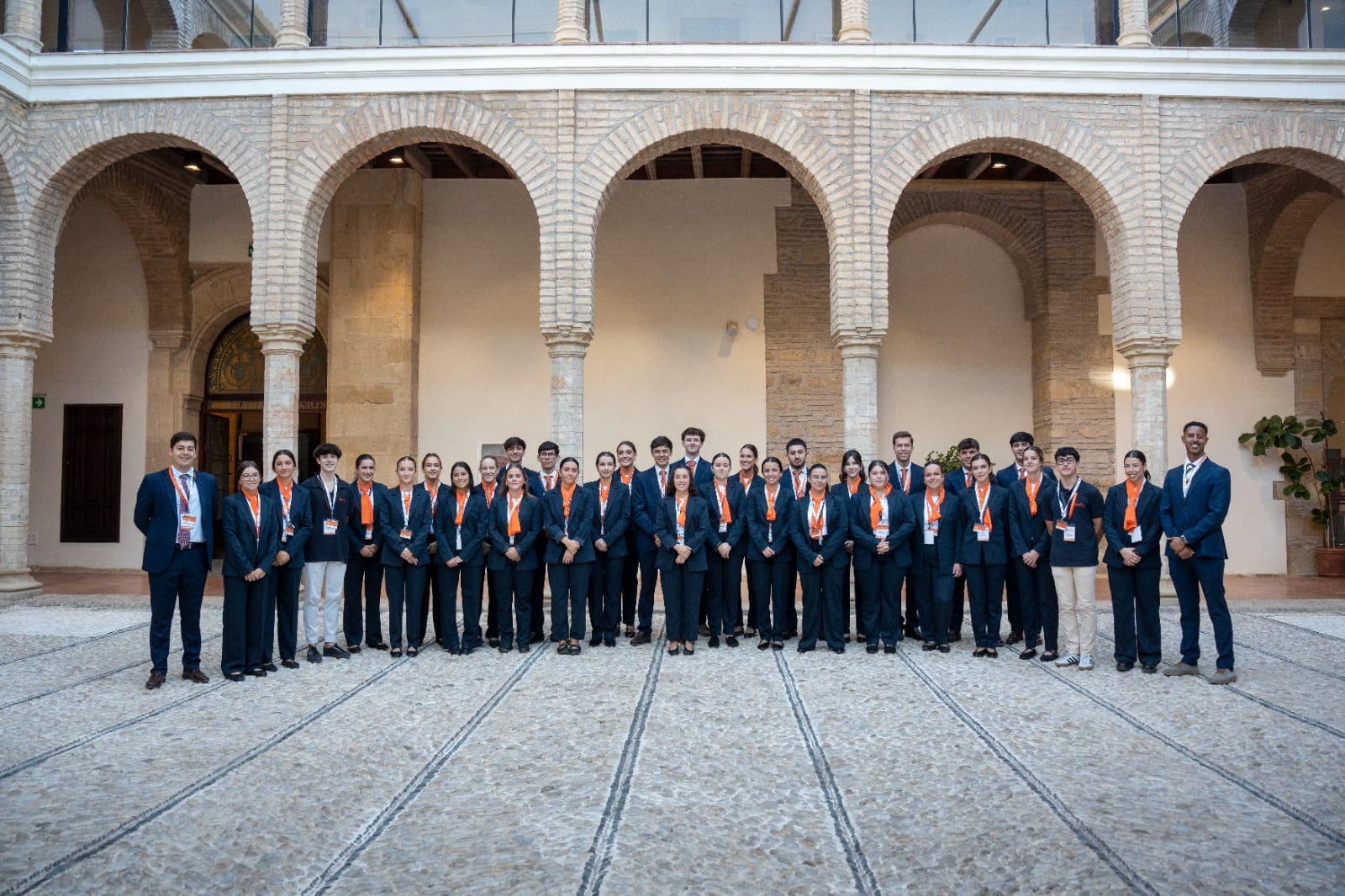 Grupo numeroso de personal de sala de Esmeeting, con traje azul y pañuelo naranja, posando en fila en el patio porticado de una sede.