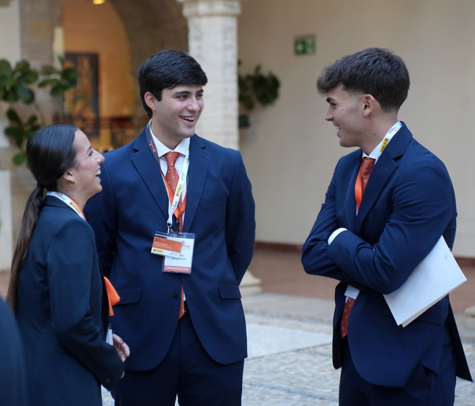 Tres integrantes del personal de sala de Esmeeting, con traje azul, conversan en un patio; uno sostiene una carpeta.