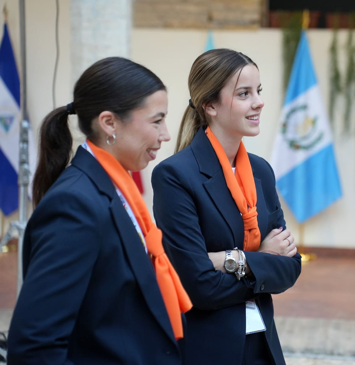 Dos integrantes del personal de sala de Esmeeting, de perfil y con pañuelo naranja, con banderas al fondo.