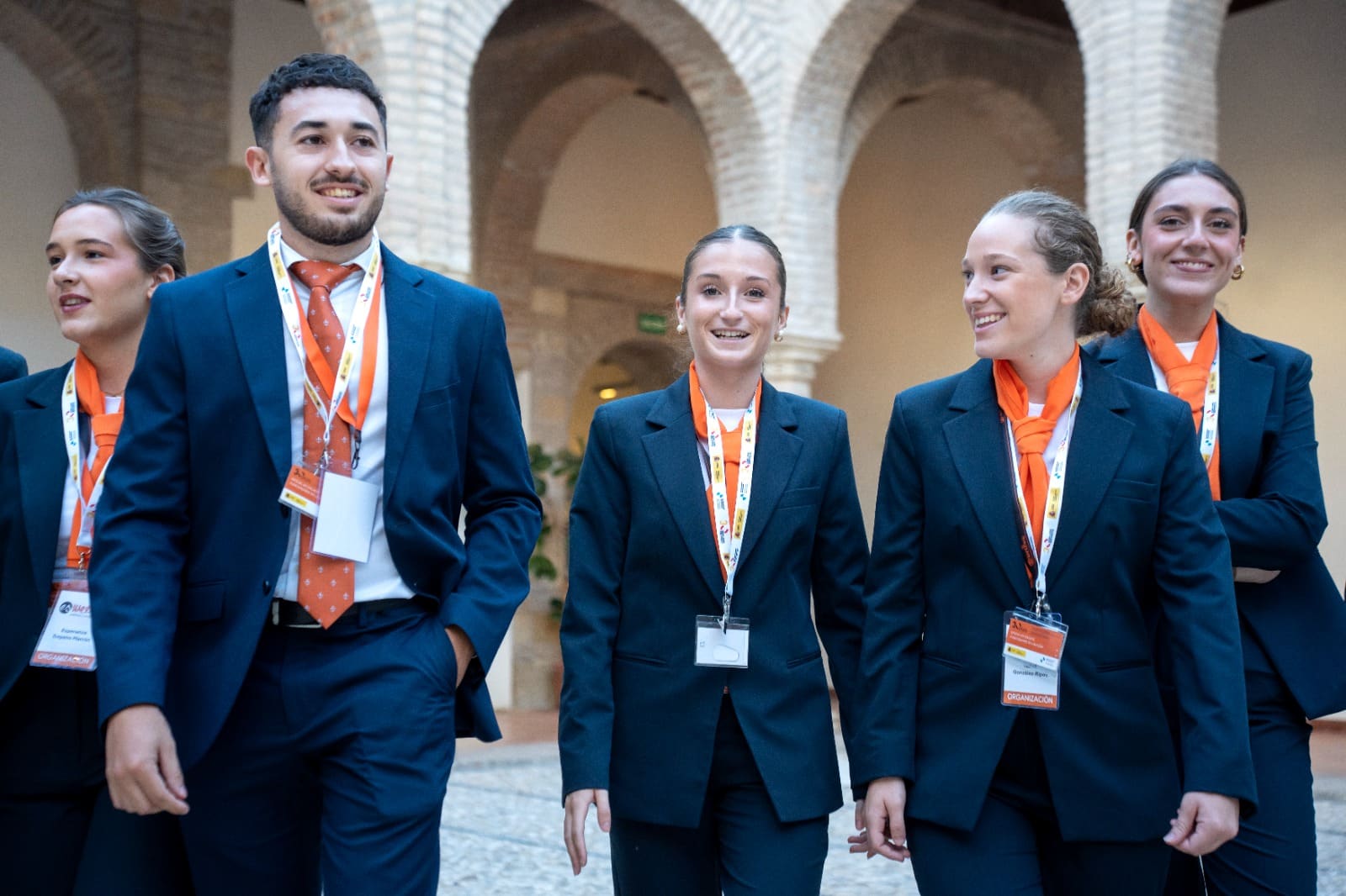 Cuatro integrantes del equipo de sala de Esmeeting, con traje azul y pañuelo naranja, caminan y sonríen durante la Cumbre Internacional.