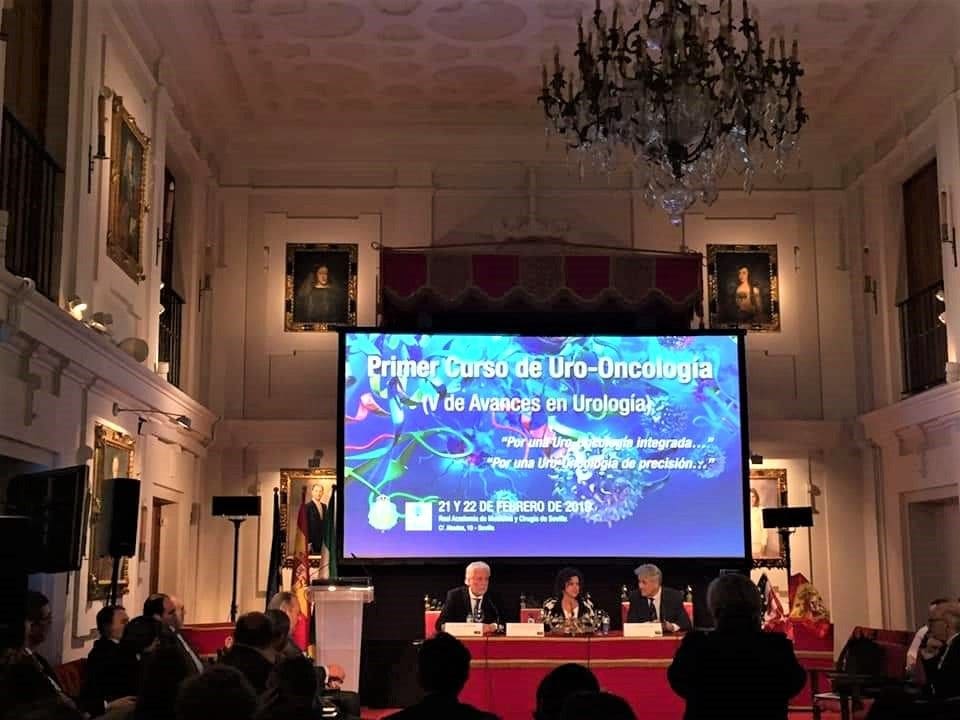 Curso de uro-oncología en un salón histórico con lámpara de araña, la mesa de ponentes y la proyección.